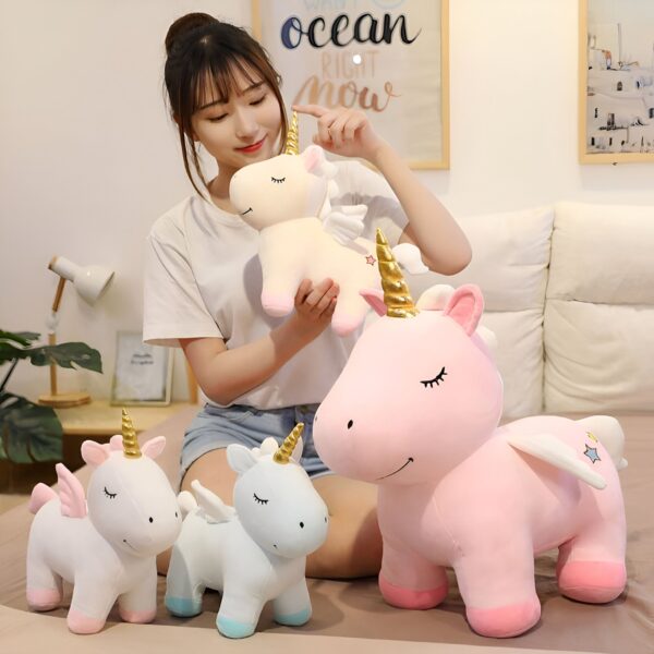 Une jeune femme tient une peluche licorne rose et blanche avec des ailes. Il y a aussi deux autres licornes en peluche sur le lit, une blanche et une bleue.