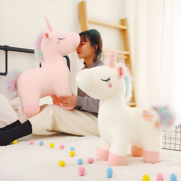 peluche-licorne-magique-douce-cadeau-enfant-4a-2 Deux peluches en forme de licorne, une rose et une blanche, avec une jeune femme assise sur un lit. La licorne rose est tenue par la jeune femme, tandis que la licorne blanche est sur le lit.