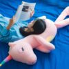Une jeune femme se blottit contre une grande peluche rose en forme de licorne tout en lisant un magazine.