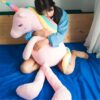 Une jeune femme assise sur un lit bleu et tenant une grande peluche licorne rose avec une crinière arc-en-ciel.