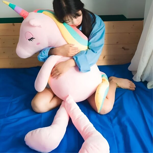 Une jeune femme assise sur un lit bleu et tenant une grande peluche licorne rose avec une crinière arc-en-ciel.
