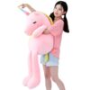 Une jeune femme souriante tient une grande peluche rose en forme de licorne avec une corne arc-en-ciel.
