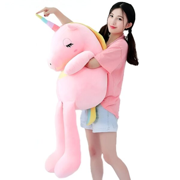 Une jeune femme souriante tient une grande peluche rose en forme de licorne avec une corne arc-en-ciel.