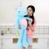 Une jeune femme tient une grande peluche licorne bleu clair avec une corne arc-en-ciel et un cœur rose.