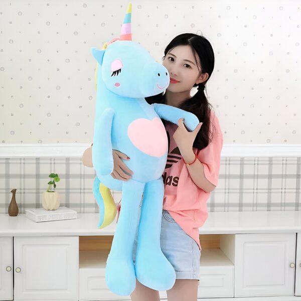 Une jeune femme tient une grande peluche licorne bleu clair avec une corne arc-en-ciel et un cœur rose.