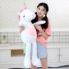 Une jeune femme souriante tenant une grande peluche de licorne blanche avec une crinière arc-en-ciel et un cœur rose.