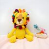 peluche-lion-tricote-colore-jouet-enfant-cadeau-unique-5a-2 Un lion en crochet jaune avec une crinière multicolore assis sur un fond rose.