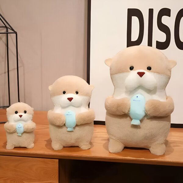 Trois peluches de loutres tenant un poisson bleu, de tailles différentes, posées sur une étagère en bois.