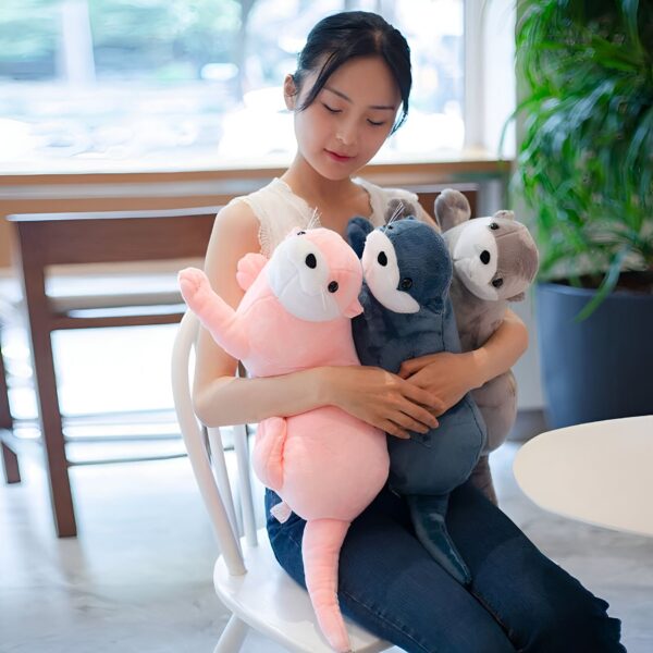 Une jeune femme est assise sur une chaise blanche et serre trois peluches en forme de loutre dans ses bras. L'une est rose, l'autre est bleu foncé et la troisième est gris clair.