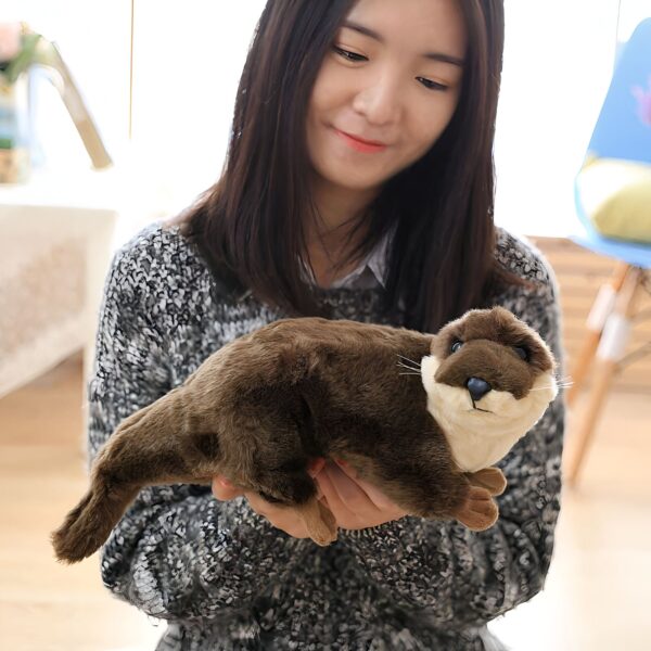 Une jeune femme tient une peluche en forme de loutre. La peluche est de couleur marron foncé et a une fourrure douce. La femme sourit et regarde la peluche avec affection.