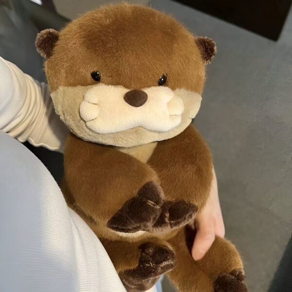 peluche-loutre-realiste-marron-3a-2 Une peluche de loutre brune en fourrure douce, avec un visage expressif et des pattes rondes. Elle est tenue dans les bras d'une personne.