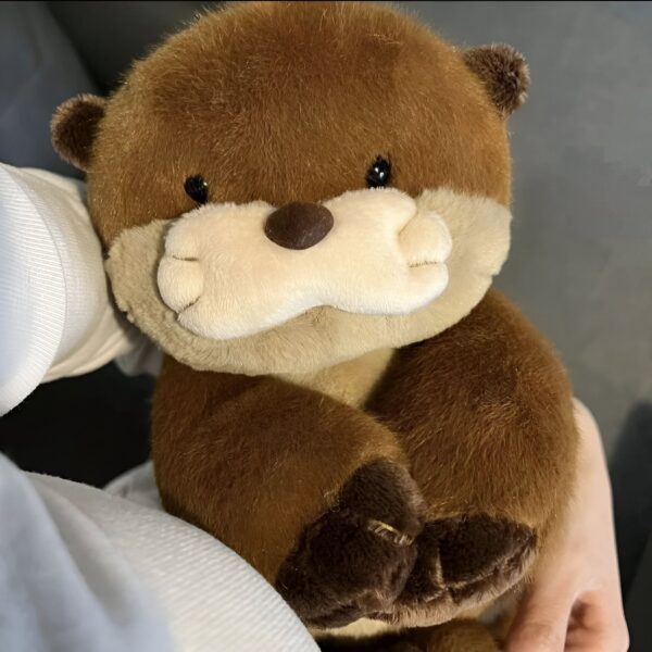 peluche-loutre-realiste-marron-5a-1 Une peluche de loutre brune et beige avec de grands yeux noirs et un petit nez. Elle est en position assise avec ses pattes repliées sur son ventre.
