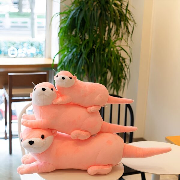 peluche-loutre-rose-jouet-doux-pour-bebe-et-enfant-cadeau-original-2a Trois peluches de loutres roses empilées sur une table. La plus grande peluche de loutre est sur le fond avec une plus petite peluche de loutre sur son dos, et la plus petite peluche de loutre est sur le dessus.
