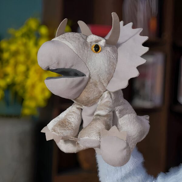 Marionnette de dinosaure Tricératops en peluche grise avec de grands yeux jaunes, portée par une main.