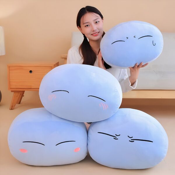 Une femme souriante tenant un coussin bleu en forme de personnage de dessin animé avec des yeux et une bouche. Il y a trois autres coussins similaires sur le canapé derrière elle.