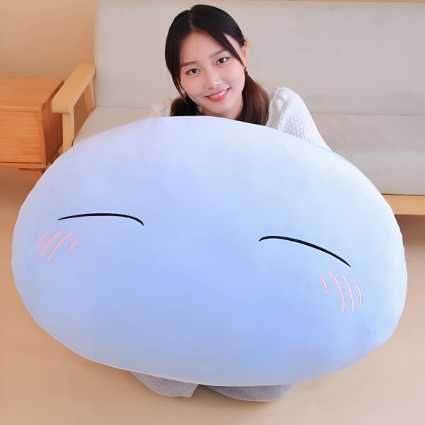Une jeune femme sourit en tenant un grand coussin bleu en forme de visage souriant avec des joues roses.