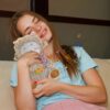 Une jeune femme aux cheveux blonds serrant un jouet en peluche en forme d'agneau et souriant, avec des notes de musique colorées flottant autour d'elle.