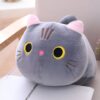 Une peluche de chat grise avec des yeux jaunes et un nez rose. Elle est posée sur un coussin blanc.