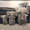 peluche-oreiller-elephant-2a Trois peluches éléphants gris, de différentes tailles, disposées sur un lit. La plus grande a une trompe rose.