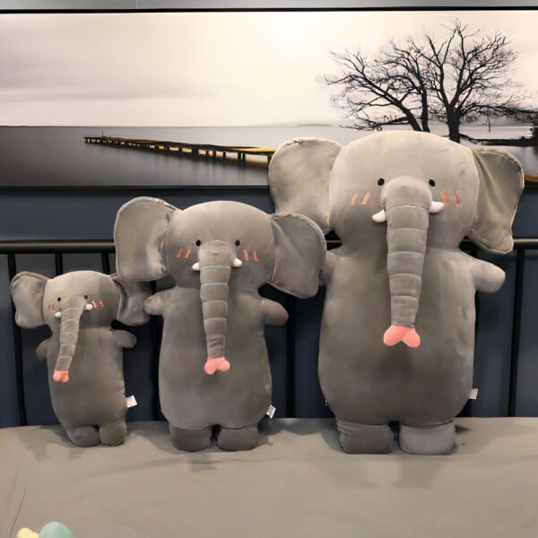 peluche-oreiller-elephant-2a Trois peluches éléphants gris, de différentes tailles, disposées sur un lit. La plus grande a une trompe rose.