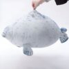peluche-oreiller-phoque-doux-et-adorable-pour-enfant-3a-1 Une peluche de phoque gris et blanc avec des points noirs, flottant dans les airs, avec une main tenant le haut de la peluche.