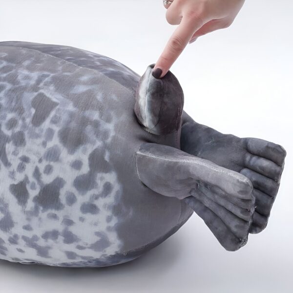 peluche-oreiller-phoque-doux-et-adorable-pour-enfant-5a-2 Une peluche de phoque gris avec des taches blanches. Un doigt pointe sur l'oreille du phoque.