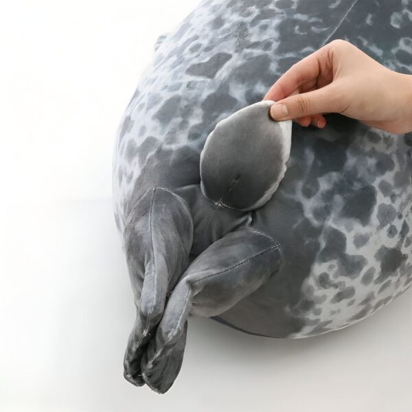Une peluche de phoque gris avec des taches blanches, une main touche la queue du phoque.