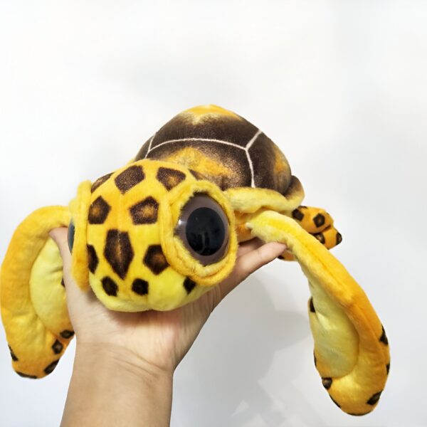 Une tortue en peluche jaune et brune avec de grands yeux noirs. La tortue est tenue dans une main.