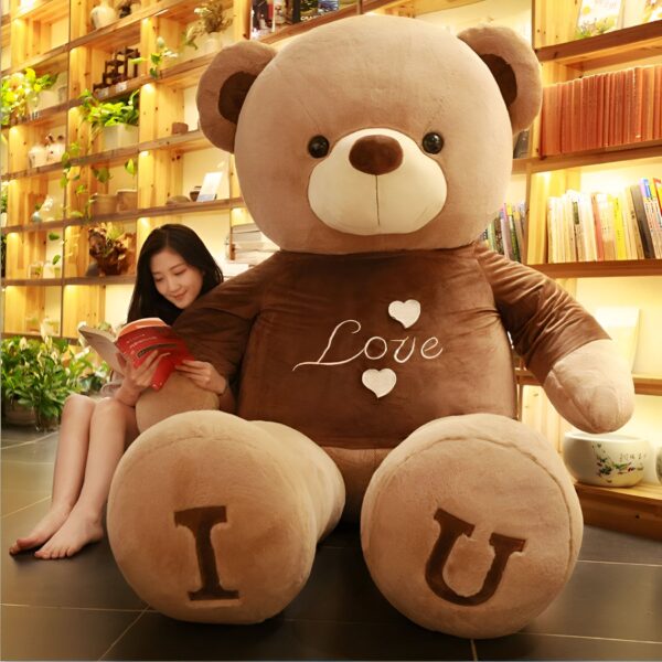 Un grand ours en peluche marron avec "Love" et "I U" écrit sur lui. L'ours est assis sur le sol devant une femme qui lit un livre.