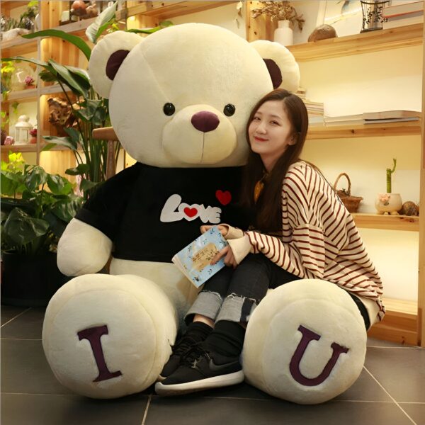 Une jeune femme souriante assise sur un grand ours en peluche blanc portant un t-shirt noir avec le mot "LOVE" et un cœur rouge. L'ours en peluche a les lettres "I" et "U" brodées sur ses pieds.