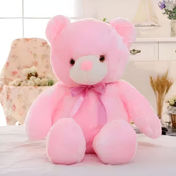 Ours en peluche rose assis sur un lit blanc avec un nœud violet autour du cou.