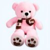 peluche-ours-rose-pleine-d-amour-1a-3 Un ours en peluche rose assis avec une écharpe brune et un cœur brun sur la poitrine. L'ours a des pattes avec des empreintes de pattes et le mot "love" sur les semelles.