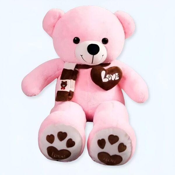 peluche-ours-rose-pleine-d-amour-1a-3 Un ours en peluche rose assis avec une écharpe brune et un cœur brun sur la poitrine. L'ours a des pattes avec des empreintes de pattes et le mot "love" sur les semelles.