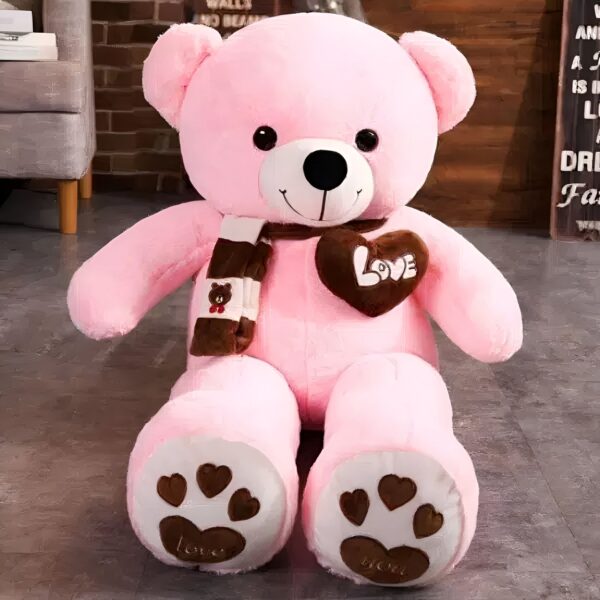 peluche-ours-rose-pleine-d-amour-2a Un grand ours en peluche rose avec un cœur marron "Love" sur la poitrine et une écharpe marron et blanche.