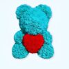 Un ours en peluche turquoise fait de roses artificielles tenant un cœur rouge.