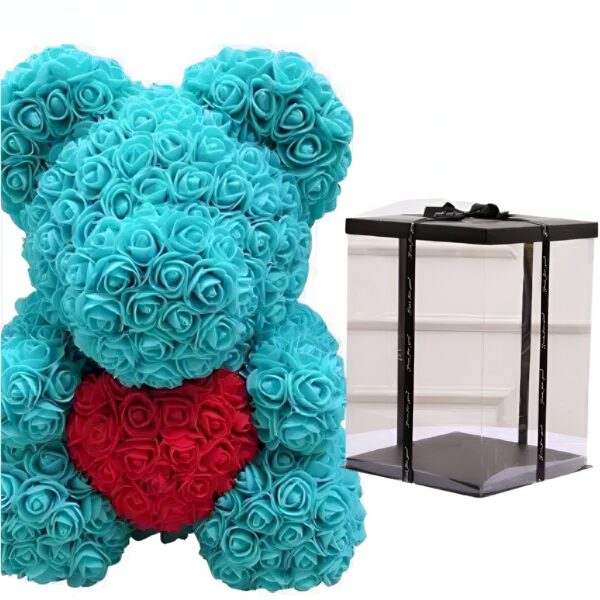 Ourson en roses artificielles de couleur turquoise tenant un cœur rouge dans ses pattes. L'ourson est emballé dans une boîte transparente avec un couvercle noir.