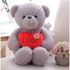 Un ours en peluche gris assis sur un fond blanc, tenant un cœur rouge avec le mot "LOVE" écrit dessus. Il y a une petite fleur rose sur le cœur. L'ours est assis sur un fond blanc avec un vase de fleurs roses à côté de lui.