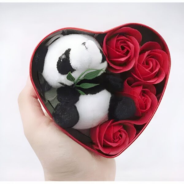 Une boîte en forme de cœur rouge contenant un petit panda en peluche blanc et noir tenant une feuille verte et entouré de roses rouges.