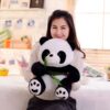 Une jeune femme souriante tient un grand ourson en peluche panda tenant une tige de bambou verte.
