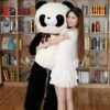 Une jeune femme souriante en robe blanche tient une grande peluche panda noire et blanche.