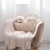 Un coussin en forme de paresseux en fourrure beige posé sur un fauteuil blanc recouvert d'un plaid beige.