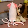 Une jeune femme souriante tient une grosse peluche rose en forme de calamar. La peluche a de grands yeux noirs et des tentacules roses.