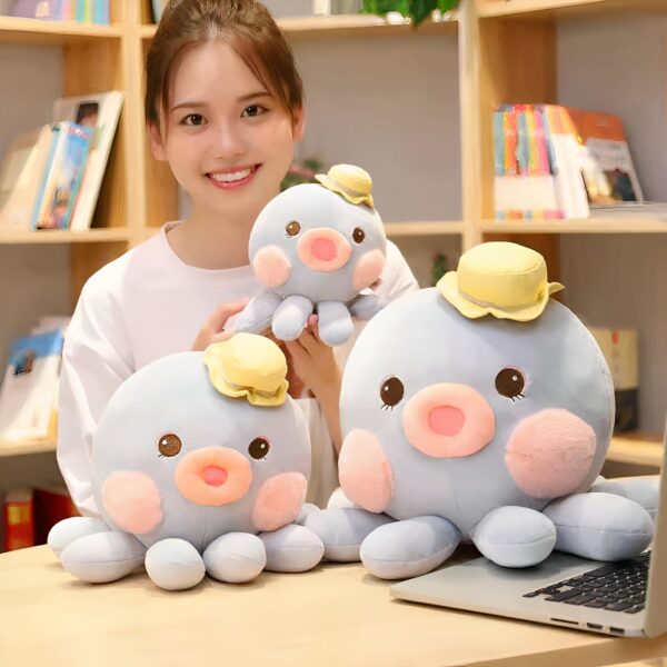 Une jeune femme souriante tient trois peluches poulpes bleu avec un chapeau jaune. Les peluches ont de grands yeux et des joues roses.