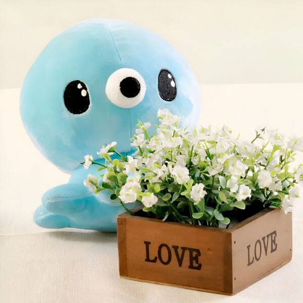Une peluche bleue en forme de phoque avec de gros yeux noirs, tenant un bouquet de petites fleurs blanches dans un pot en bois avec l'inscription "LOVE" gravée sur deux côtés.