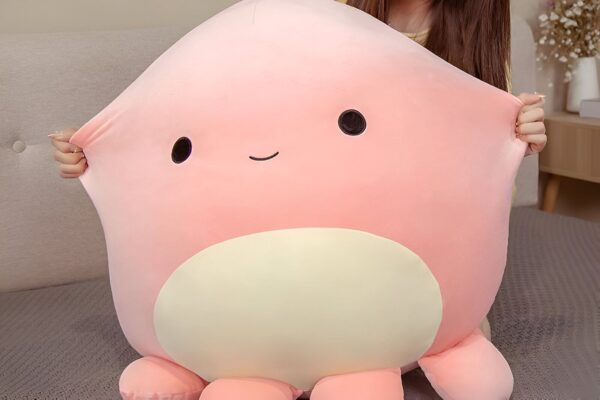 peluche-pieuvre-oreiller-doux-10a-1 Une grande peluche rose en forme de poulpe avec un grand sourire et des yeux noirs.