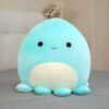 peluche-pieuvre-oreiller-doux-1a-1 Une peluche en forme de pieuvre bleu avec un grand sourire, posée sur un lit gris.