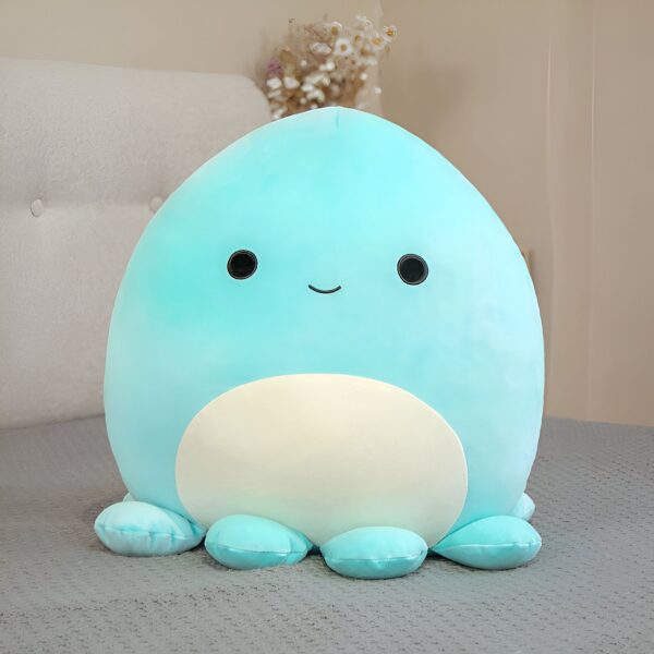 peluche-pieuvre-oreiller-doux-1a-1 Une peluche en forme de pieuvre bleu avec un grand sourire, posée sur un lit gris.