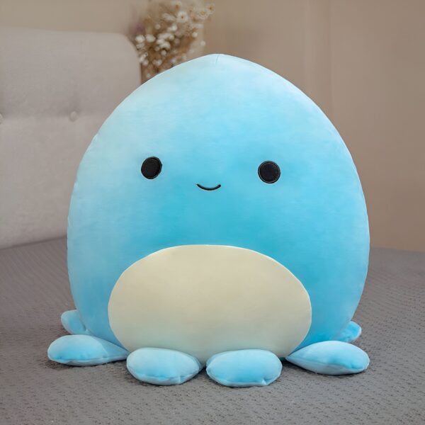 peluche-pieuvre-oreiller-doux-5a-6 Une peluche bleue en forme de pieuvre avec un sourire et des yeux noirs. Elle est posée sur un lit gris.