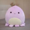 peluche-pieuvre-oreiller-doux-6a-5 Une peluche violette en forme de poulpe souriant avec des yeux noirs, posée sur un lit gris.