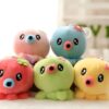 Cinq adorables peluches poulpes en rose, bleu, rouge, vert et jaune avec des yeux noirs et une bouche rose. Ils portent tous des fleurs colorées sur la tête.
